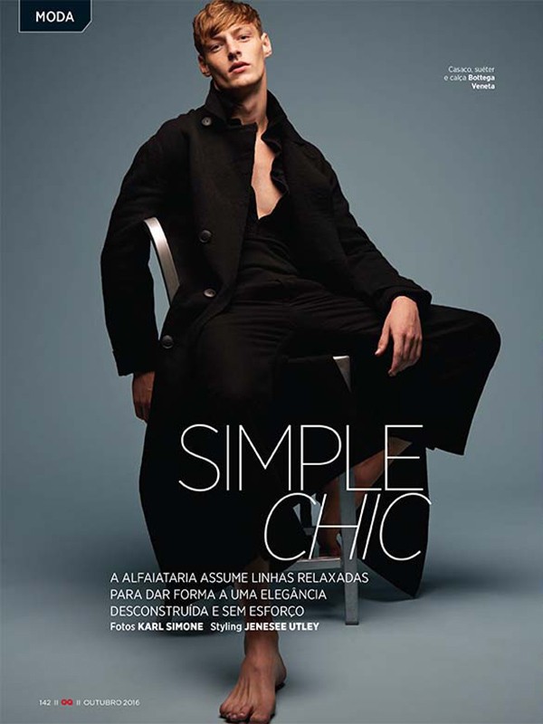 A light-skinned male model in a black outfit, sitting on a metal chair in a studio setting, is featured in a fashion editorial shot at eye-level.