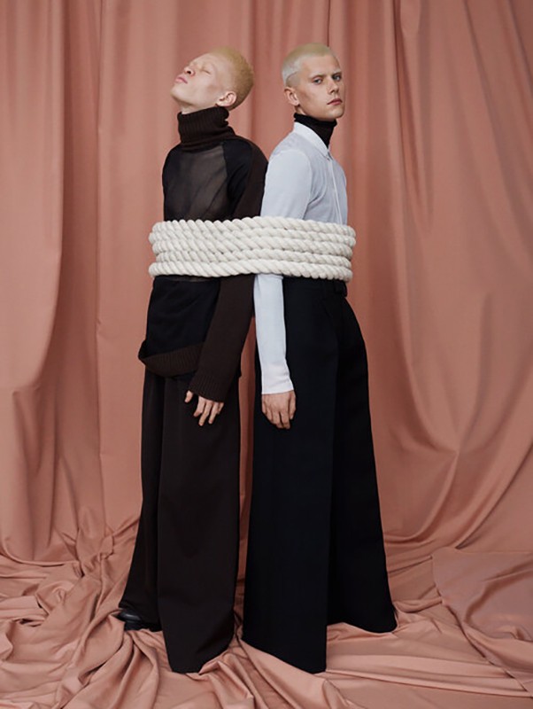 Two fair-skinned adults pose in black and white high-fashion clothing. A thick rope is wrapped around them, set against a salmon-colored backdrop.
