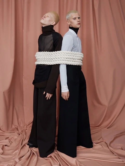Two fair-skinned adults pose in black and white high-fashion clothing. A thick rope is wrapped around them, set against a salmon-colored backdrop.