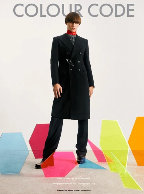 Fashion model in a black coat, standing indoors on a light-colored floor among colorful geometric acrylic glass objects, fashion editorial.