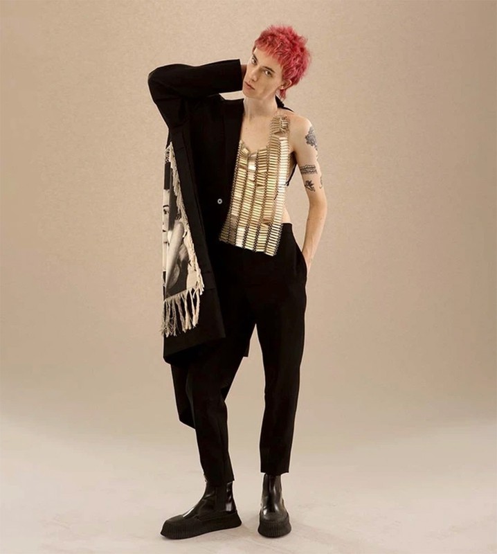 A person with dyed pink hair in an oversized black blazer with a decorative scarf, a metallic top, black pants and black boots.