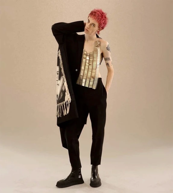 A person with dyed pink hair in an oversized black blazer with a decorative scarf, a metallic top, black pants and black boots.