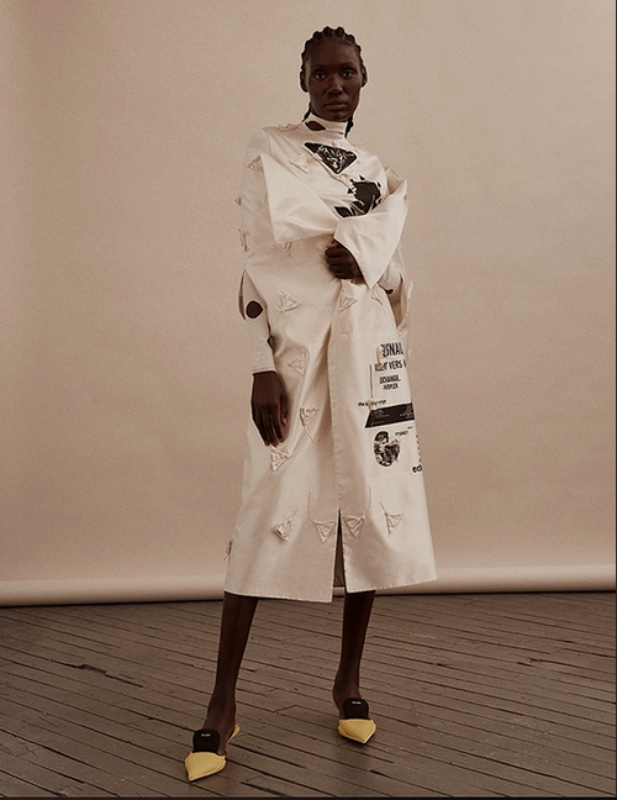 Full-length portrait of a slender, dark-skinned female fashion model in a long white designer dress with yellow heels, standing indoors.