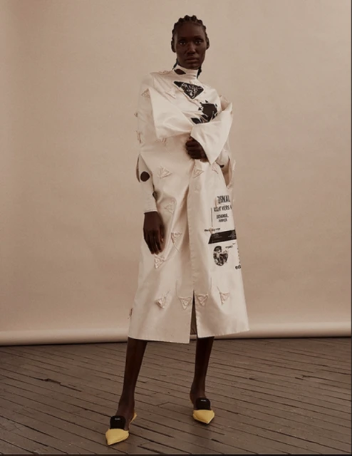 Full-length portrait of a slender, dark-skinned female fashion model in a long white designer dress with yellow heels, standing indoors.