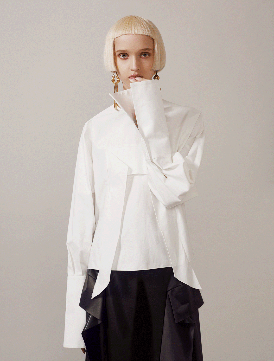 A model poses in a white long-sleeved collared shirt and black skirt in a studio with a neutral background for a fashion editorial shoot.