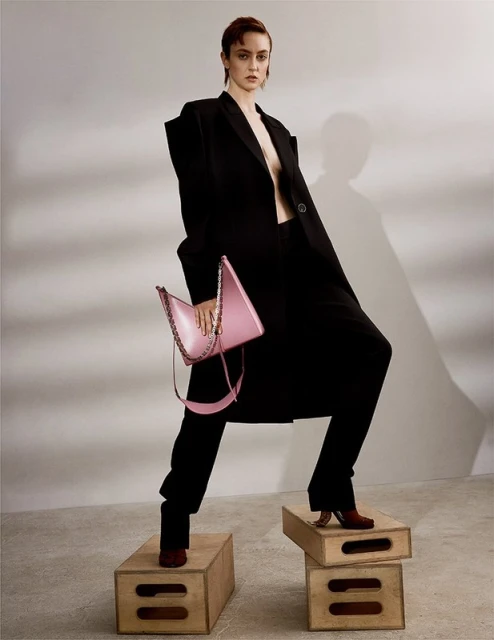 Fashion editorial shot of a stylish woman in a black coat standing on boxes holding a pink handbag in a neutral coloured studio.
