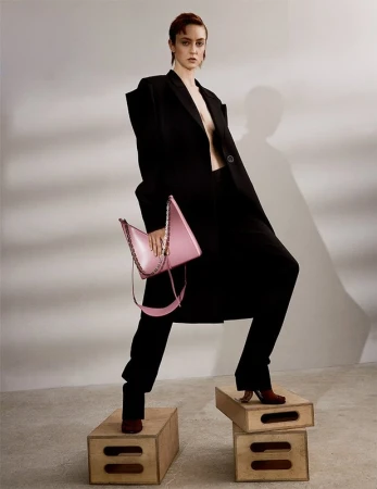 Fashion editorial shot of a stylish woman in a black coat standing on boxes holding a pink handbag in a neutral coloured studio.