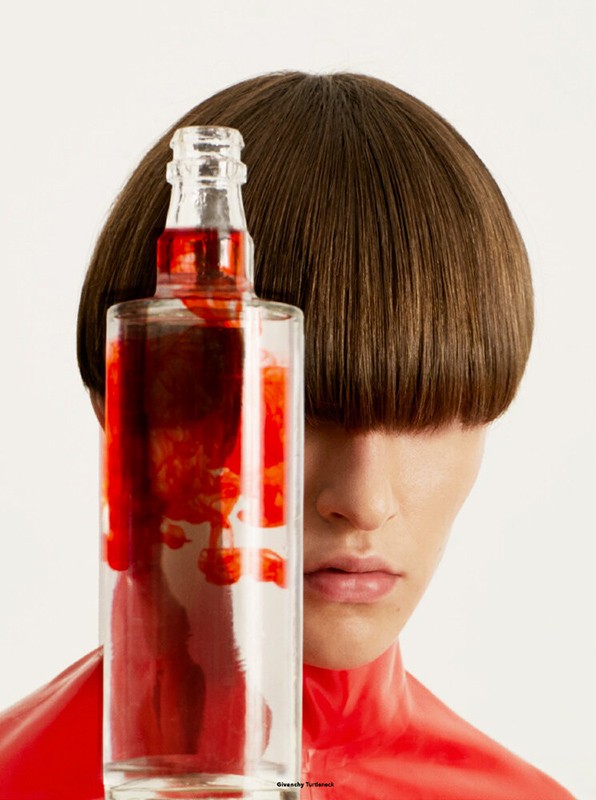Person with a modern bowl haircut stands behind a glass bottle containing a red liquid, set against a plain white backdrop.