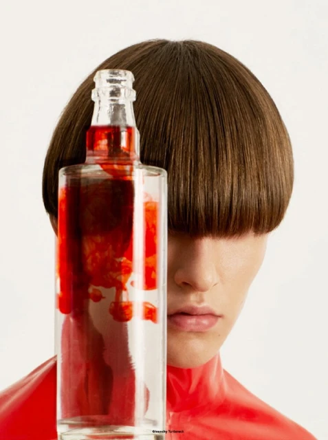 Person with a modern bowl haircut stands behind a glass bottle containing a red liquid, set against a plain white backdrop.