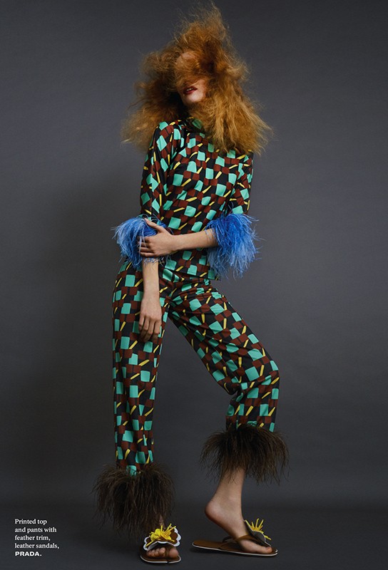 Fashion editorial shot of a woman in a patterned top and pants with feather trim, posing against a neutral gray backdrop in studio lighting.