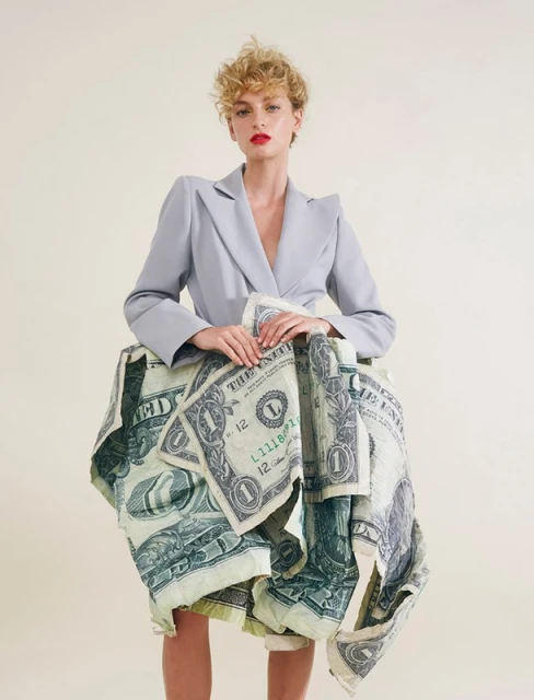 Adult female model posing in a grey blazer and a skirt constructed of US one-dollar bills against a light beige studio backdrop.