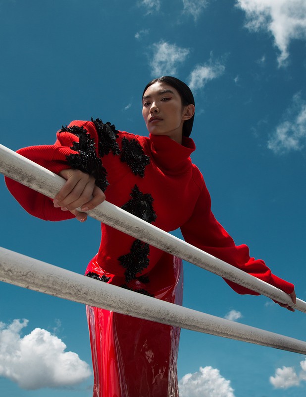 Fashionable Asian woman in a red outfit poses on a railing against a blue sky with clouds, showcasing a blend of elegance and style.