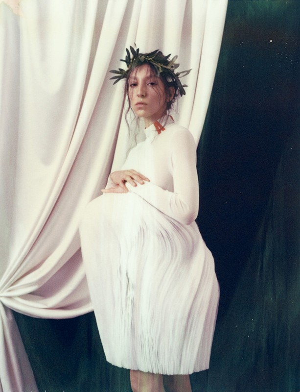 An adult woman wears a white dress and a leafy crown while posing in front of draped white and dark-green fabric in a studio setting.