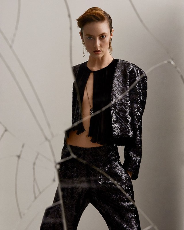 A stylish adult female model wearing a black sequined jacket and pants poses in a photo studio setting with a broken glass foreground.
