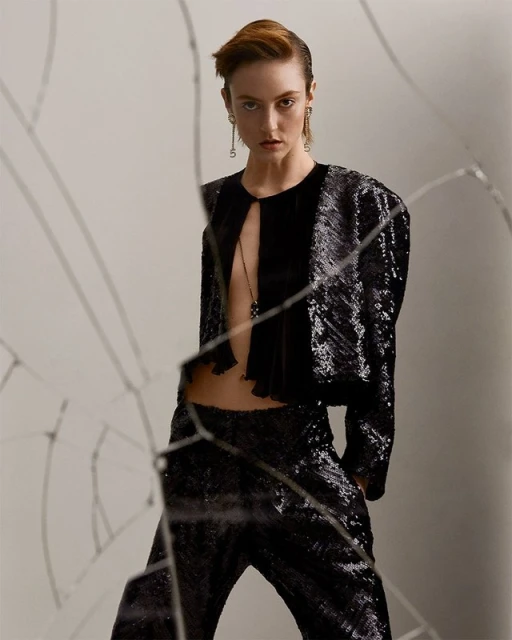 A stylish adult female model wearing a black sequined jacket and pants poses in a photo studio setting with a broken glass foreground.