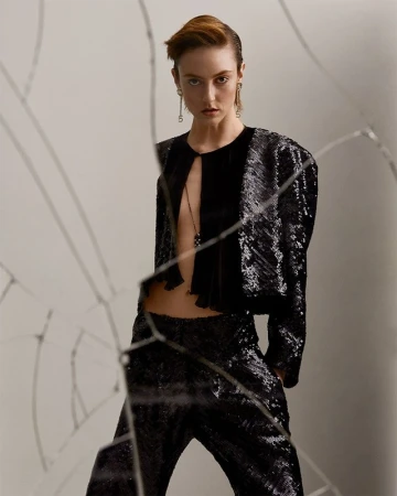 A stylish adult female model wearing a black sequined jacket and pants poses in a photo studio setting with a broken glass foreground.
