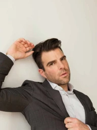 Zachary Quinto in a grey suit jacket posing against a plain neutral wall
