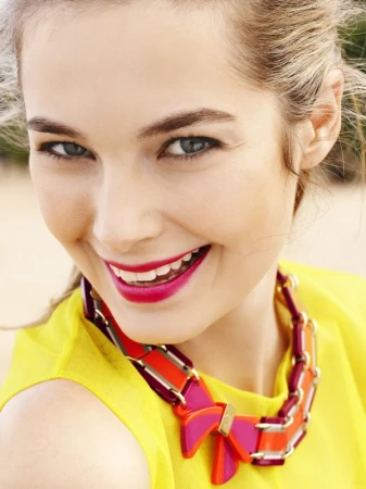 Close-up portrait of a woman with bright pink lipstick wearing a yellow top and a colourful chunky statement necklace.