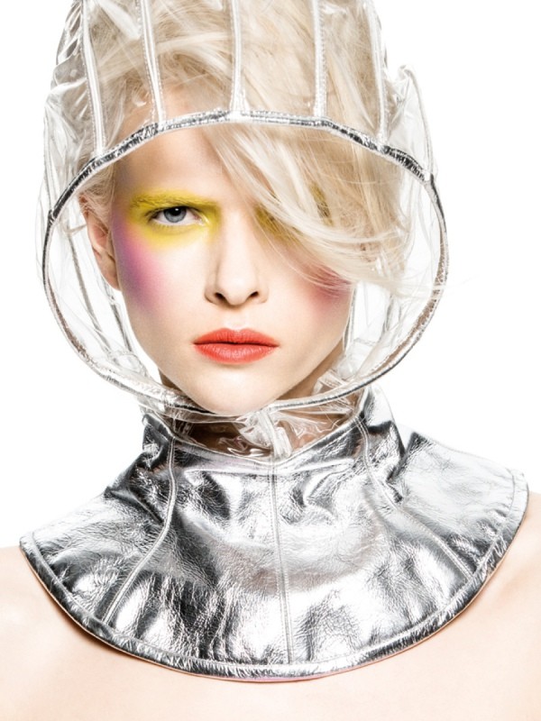Woman with pale blonde hair wears futuristic clear vinyl hood and metallic collar with yellow and pink eyeshadow in a studio shot.