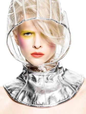 Woman with pale blonde hair wears futuristic clear vinyl hood and metallic collar with yellow and pink eyeshadow in a studio shot.