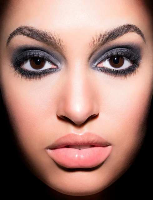 Close-up portrait of a woman with smokey eyeshadow, featuring brown eyes, full pink lips, and smooth skin against a dark background.