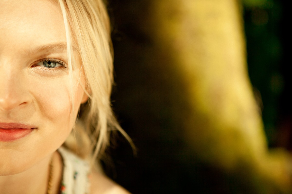 Close-up portrait of a smiling blonde woman with blue eyes, partially visible, against a blurred outdoor background with yellow lighting.