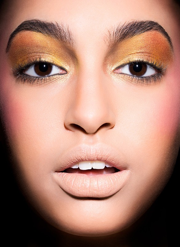 Close-up portrait of a woman with gold eyeshadow and beige lipstick, facing the camera with a neutral expression against a dark backdrop.