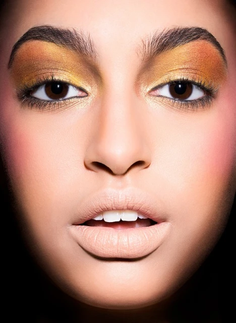 Close-up portrait of a woman with gold eyeshadow and beige lipstick, facing the camera with a neutral expression against a dark backdrop.