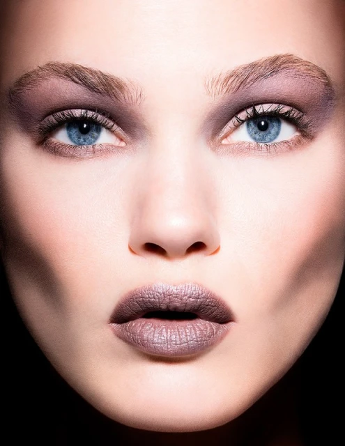 Close-up portrait of a woman with fair skin, blue eyes, and mauve-toned eye shadow and lipstick against a dark background in studio lighting.