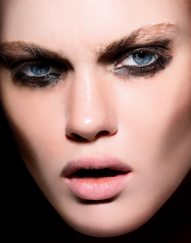 Close-up portrait of a fair-skinned woman with blue eyes wearing heavy black eye makeup, capturing her intense gaze in dark lighting.