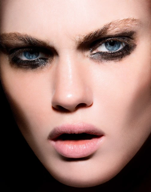 Close-up portrait of a fair-skinned woman with blue eyes wearing heavy black eye makeup, capturing her intense gaze in dark lighting.