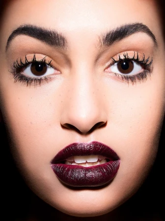 Close-up shot of a woman's face featuring prominent eyes with long lashes and dark plum lipstick, set against a dark background.