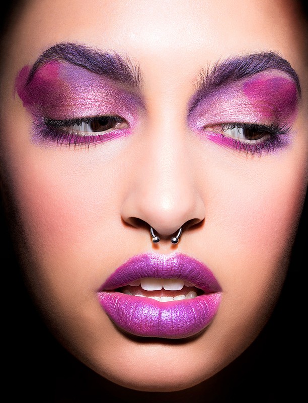 Close-up of a woman's face with dramatic purple makeup on her eyebrows, eyelids, and lips, set against a stark black background.