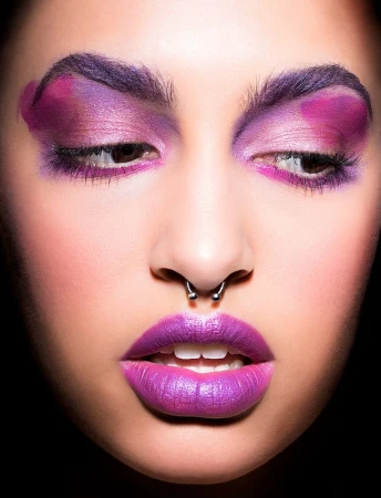 Close-up of a woman's face with dramatic purple makeup on her eyebrows, eyelids, and lips, set against a stark black background.