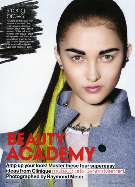 Portrait of a young woman with yellow streaks in dark hair, heavy brow makeup, wearing gray coat and spike necklace, on a magazine page.