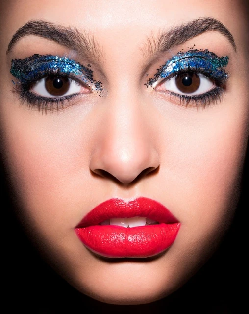 Close-up portrait of a woman with glamorous blue glitter eyeshadow and bright red lipstick against a dark background.