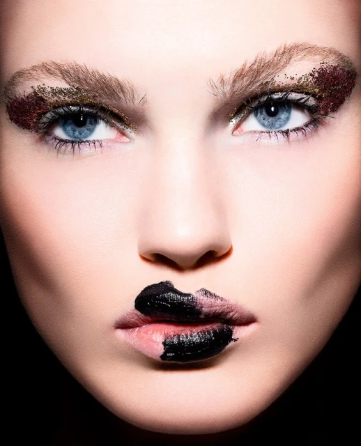 Close-up portrait of a woman with striking blue eyes, glitter eye makeup, and partially black-painted lips against a dark background.