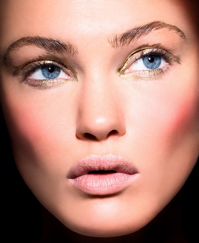 Close-up of a woman's face with striking blue eyes, gold eyeshadow, and fair skin, set against a dark background. Beauty shot, beauty concept.