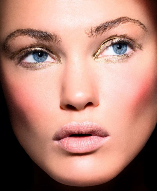 Close-up of a woman's face with striking blue eyes, gold eyeshadow, and fair skin, set against a dark background. Beauty shot, beauty concept.