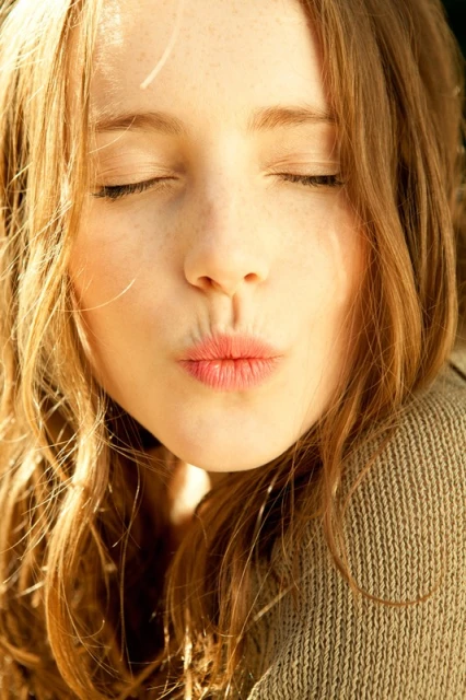 Close-up portrait of a young adult female with fair skin and wavy red hair, eyes closed, puckering her lips in a kiss, wearing a knit sweater.