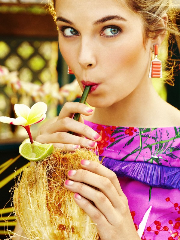 Attractive fair-skinned woman drinks tropical beverage through a straw, adorned with a flower and lime, pink nails, and a floral dress.