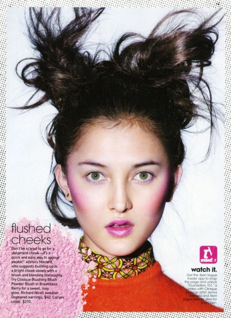 Close-up portrait of a young woman with updo hairstyle, wearing makeup and a patterned top, against a halftone background.