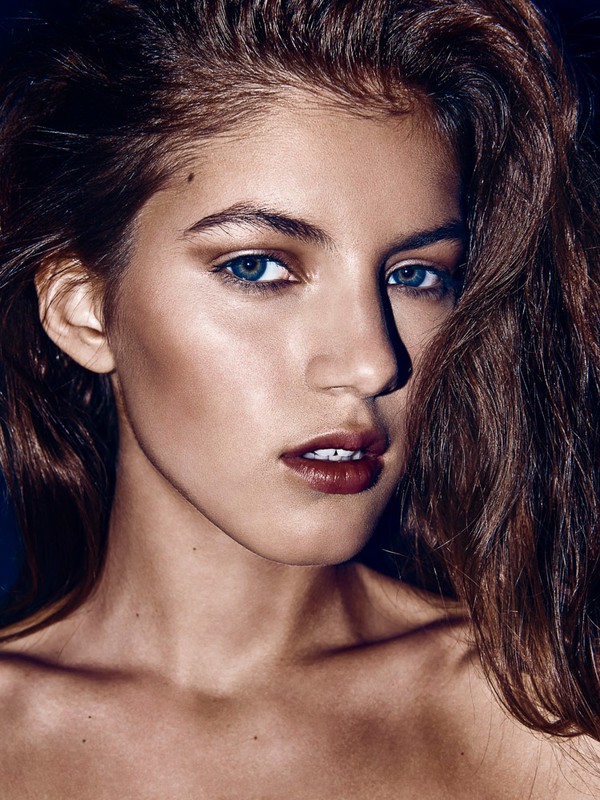 Close-up portrait of a woman with long brown hair, blue eyes, and dark lipstick, styled by Jenna Menard.