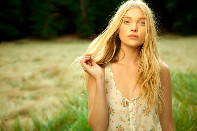 Blonde woman with long hair wearing a floral top posing in an outdoor meadow with green grass.