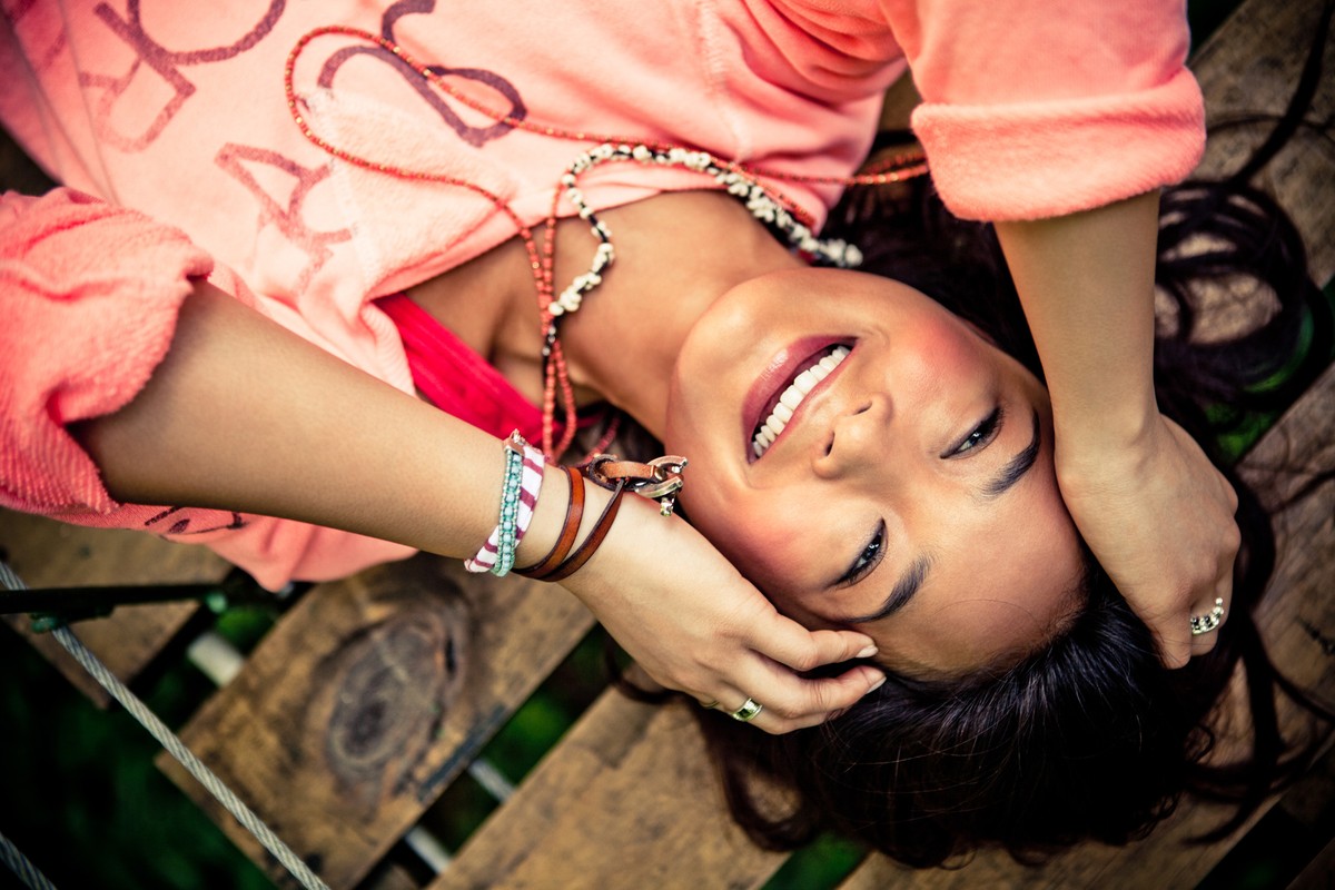 Smiling woman with her hands behind her head laying on a wooden surface wearing a peach sweatshirt with colorful bracelets and necklaces.