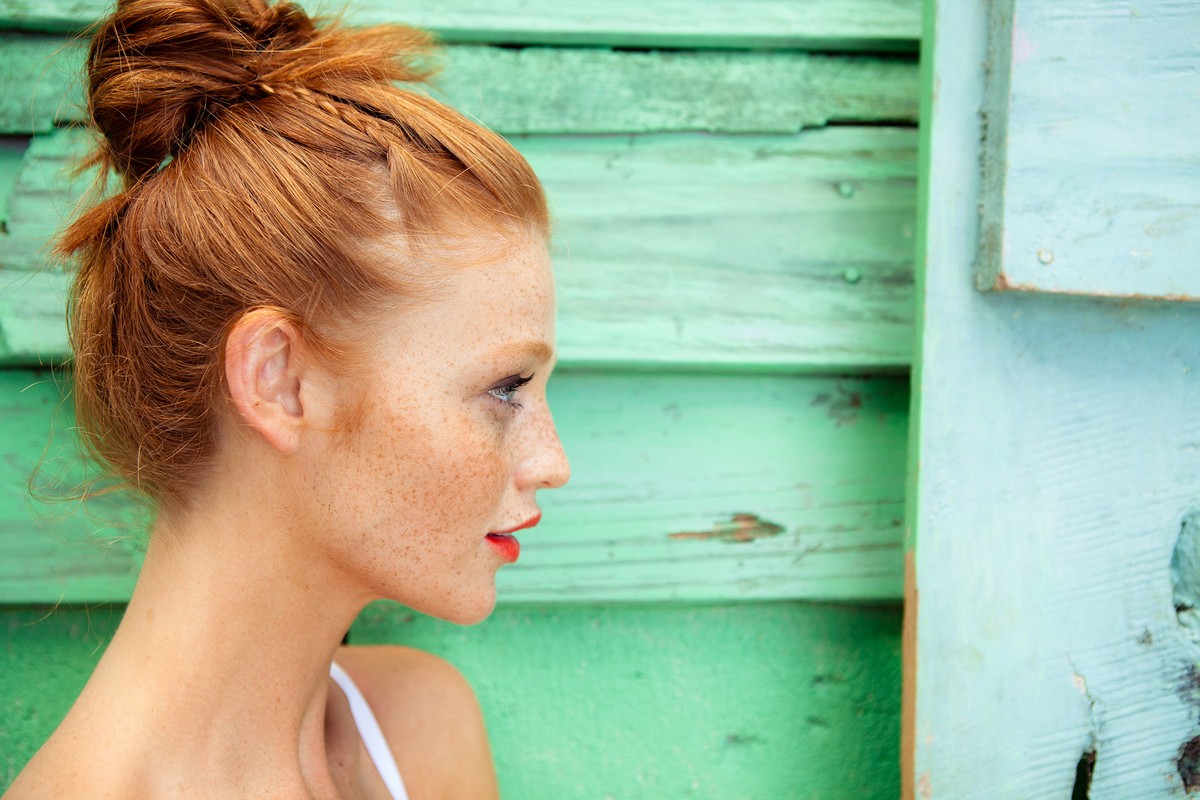 Close-up of a red-haired woman in profile with an updo, facing right, posing in front of a light green and blue distressed wood panel.