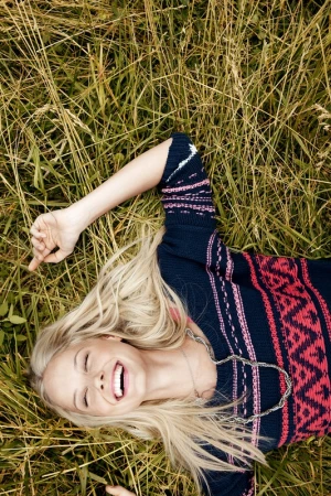 Blonde woman laughing while lying in tall green grass wearing a patterned navy blue knit sweater with pink accents.