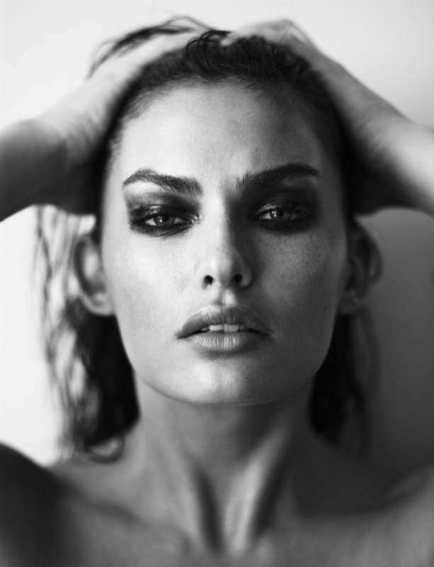 Close-up portrait of a woman in black and white with dark eye makeup holding her wet hair with both hands.