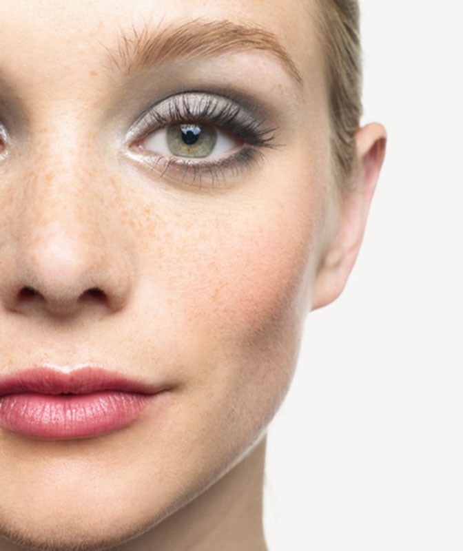 Close-up of a young woman's face with fair skin, visible freckles, and green eyes. She has dramatic grey smokey eye makeup and glossy lips.