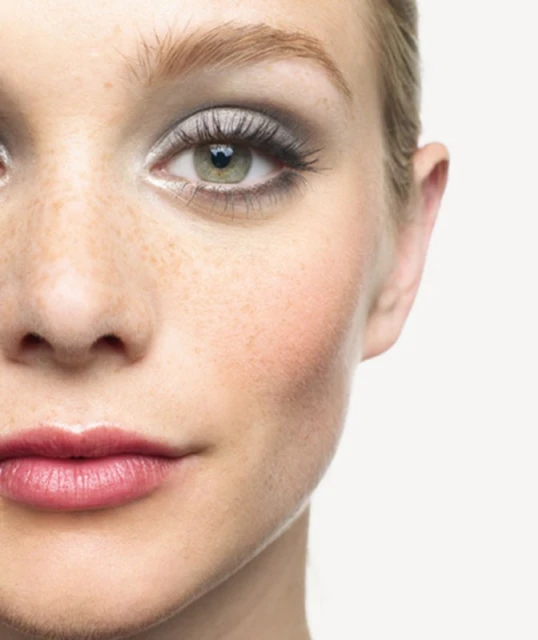 Close-up of a young woman's face with fair skin, visible freckles, and green eyes. She has dramatic grey smokey eye makeup and glossy lips.